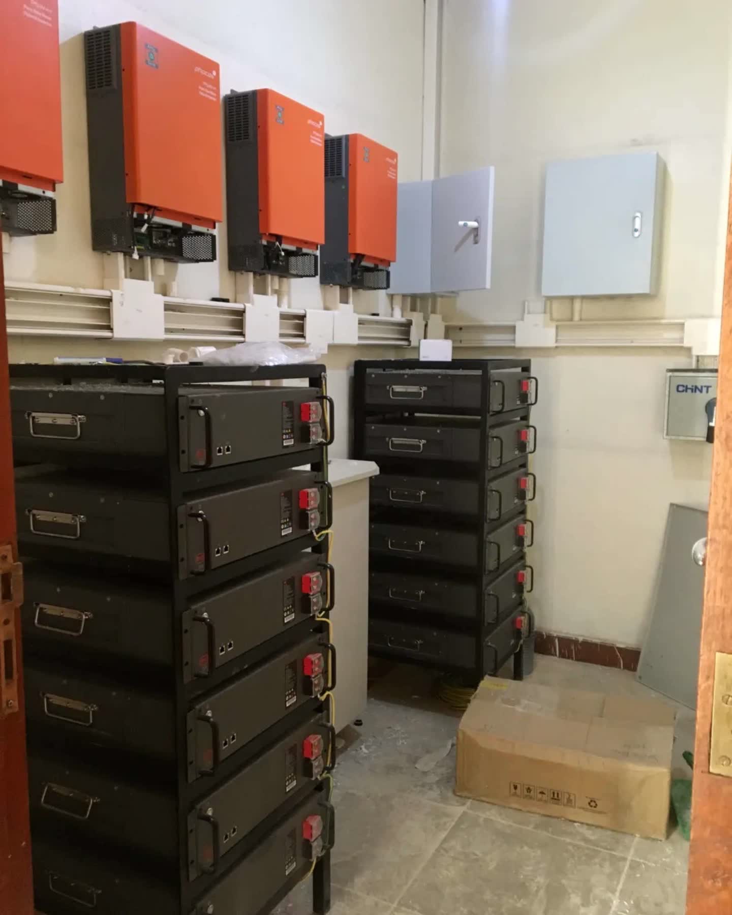 A battery room with three wall mounted inverters above two racks of lithium battery modules and distribution boards.