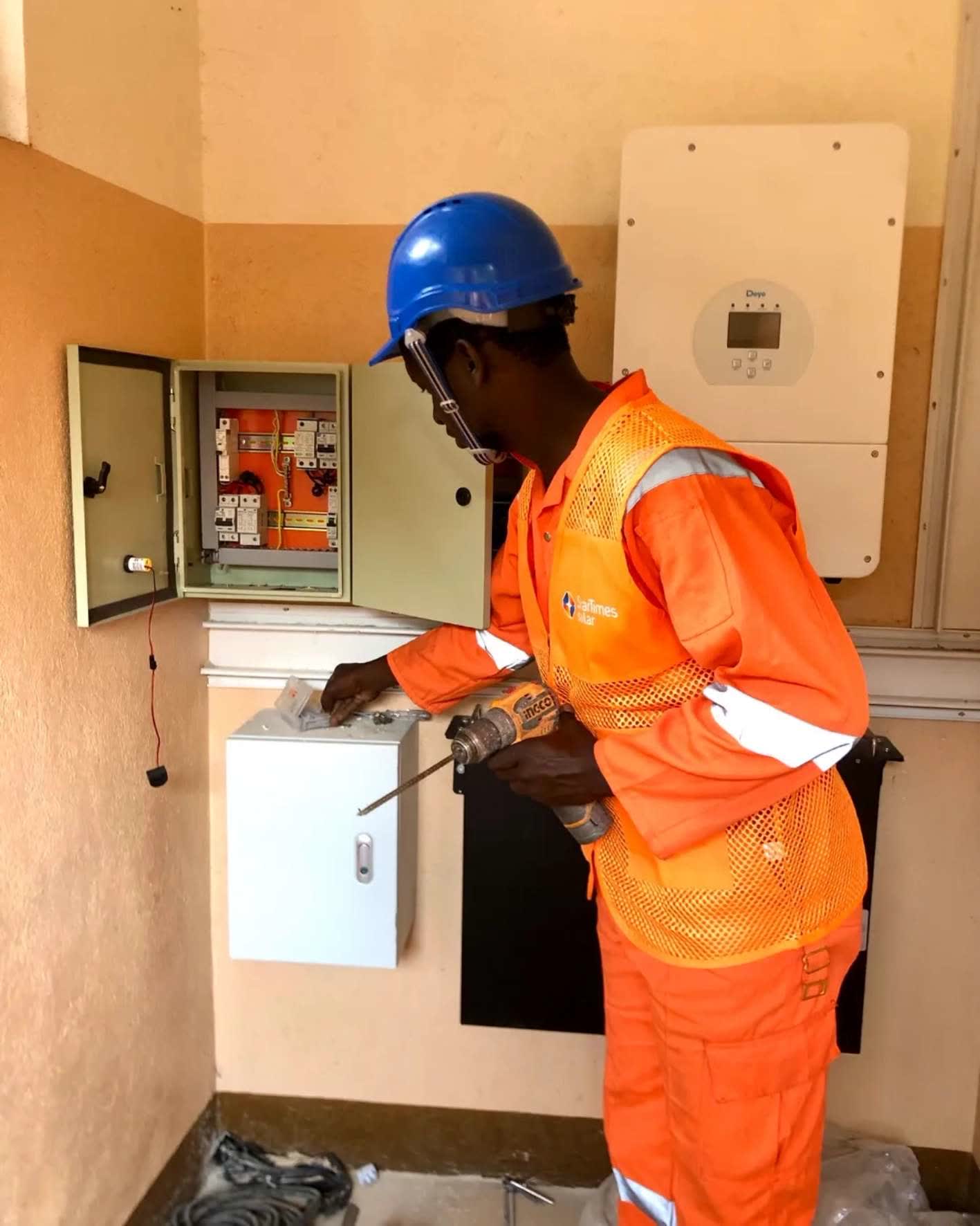 A Bakaluba engineer in orange protective clothing fitting a distribution board next to a wall mounted solar inverter.