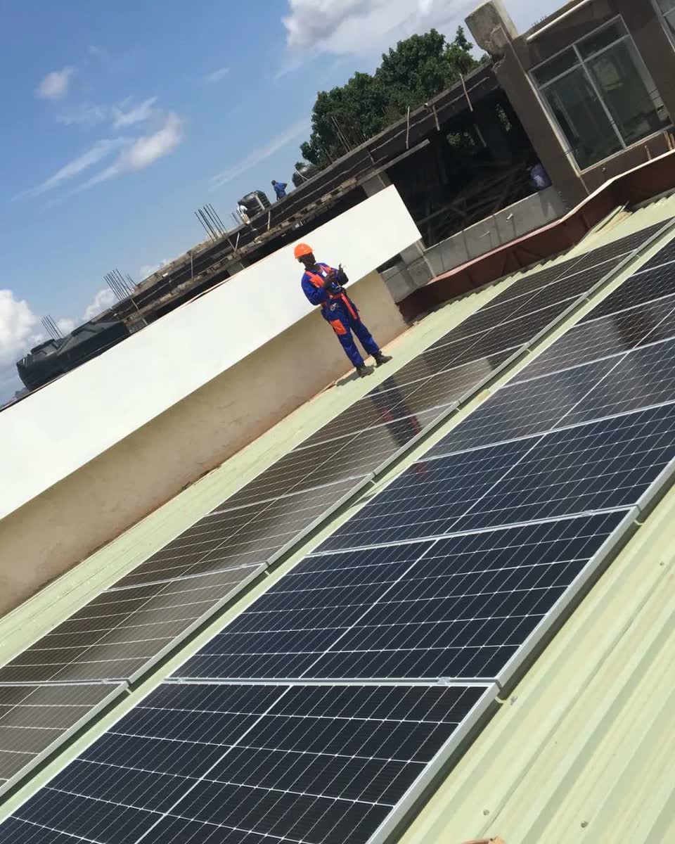 A Bakaluba technician in a hard hat standing on a metal roof beside a newly installed solar array.